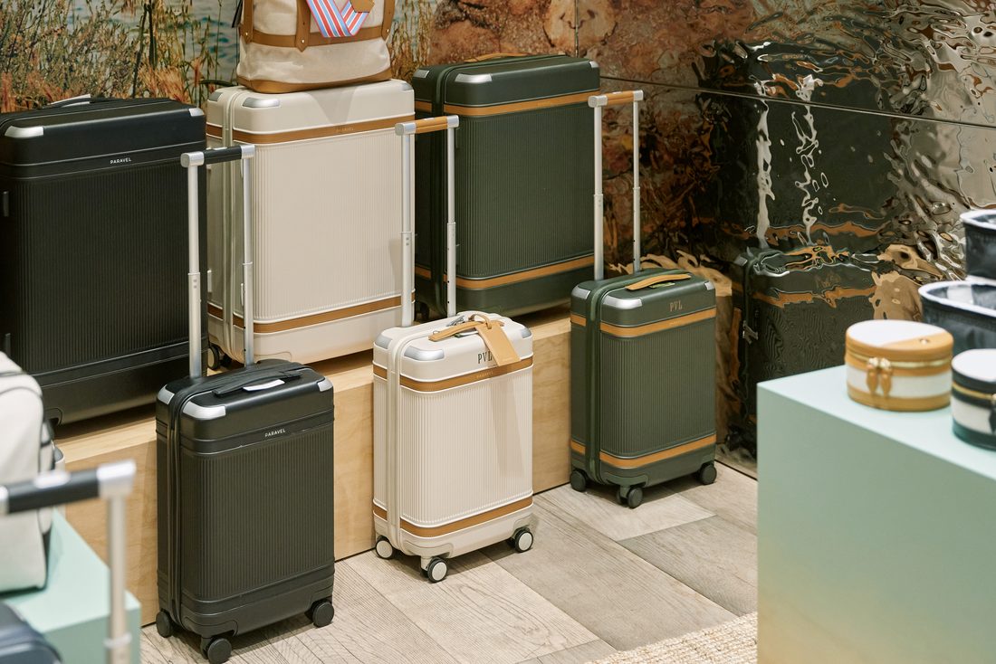 Black, olive-green, and cream hardside Aviator cases, monogrammed PVL, clustered on low maple plinths against the cliff mural and a rippled mirror wall, with round vanity cases on a mint plinth at right.