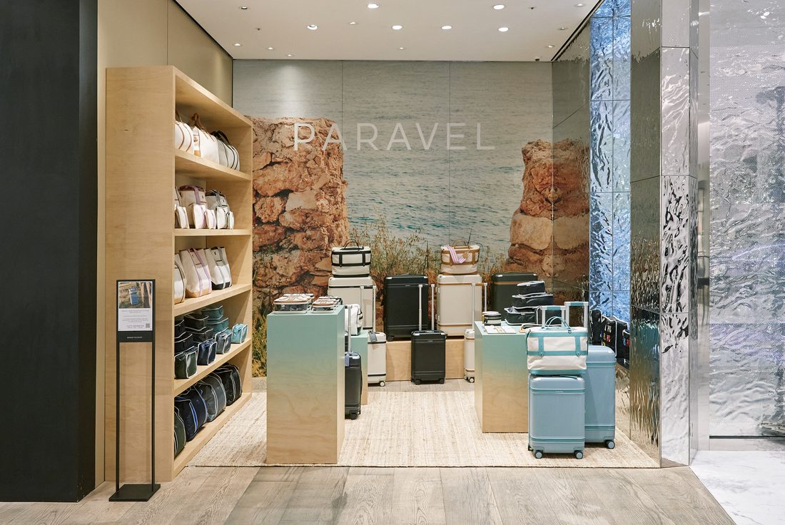 The Paravel shop-in-shop at Boon the Shop: maple shelving of packing cubes at left, a coastal-cliff mural behind the PARAVEL wordmark, and pale-blue Aviator cases on maple plinths.