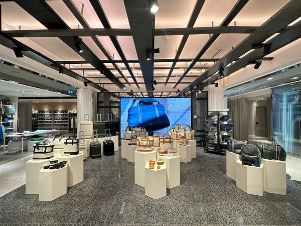 A wider view of the Doha floor: a black grid ceiling and terrazzo floor with white faceted plinths of luggage and a screen showing a blue duffel.
