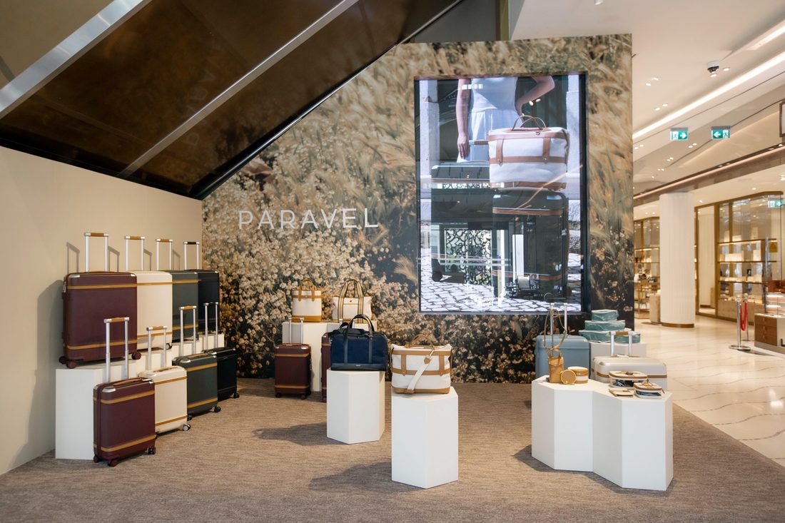 The Paravel space at Galeries Lafayette Doha: a wall printed with a dried-floral field behind the PARAVEL wordmark, a tall screen playing the brand film, and rolling luggage with white faceted plinths of totes and bags.