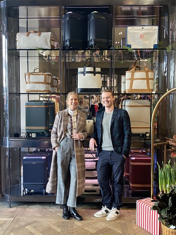 A glass-fronted display cabinet of Paravel luggage in black, navy, cream, and lilac at the Madison Avenue flagship, with two people standing in front amid holiday greenery.