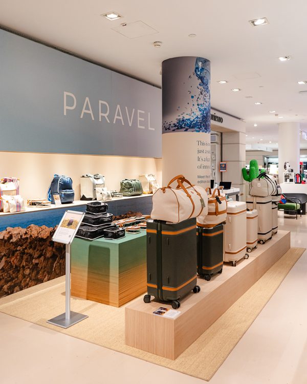The Paravel corner at La Rinascente Milan: a pale-blue PARAVEL wall, a column wrapped in a blue water-splash print, and a maple riser lined with green-and-cream Aviator luggage.