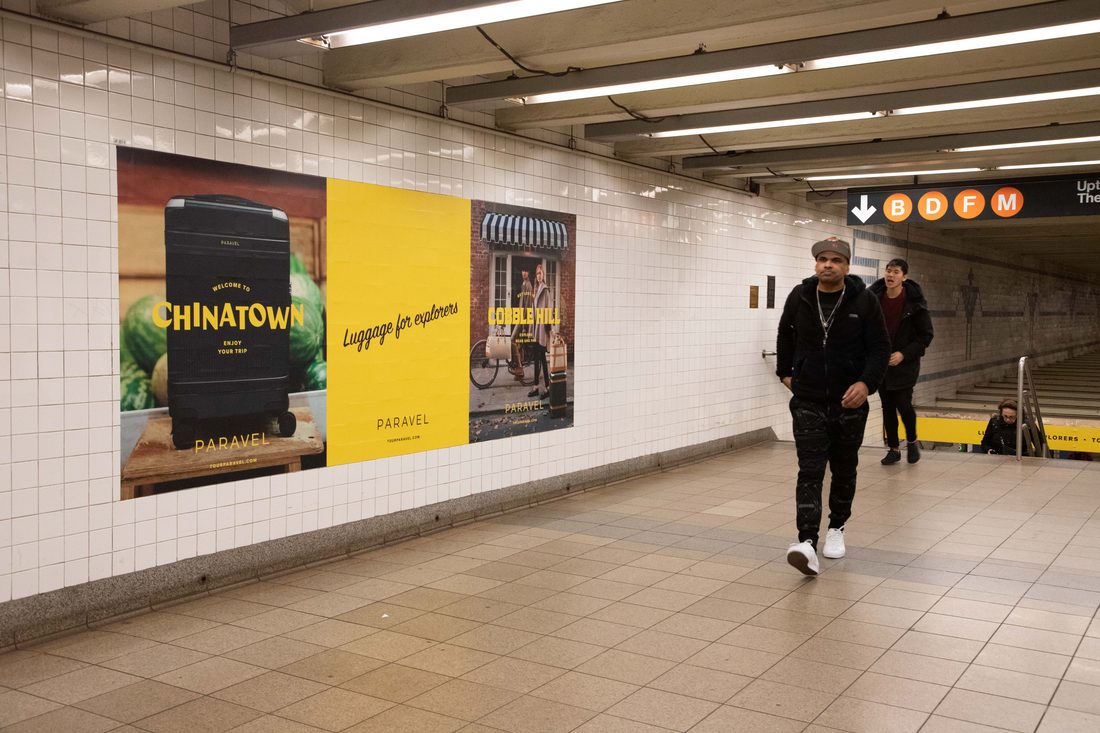 Commuters walking the white-tiled Broadway-Lafayette passage past PARAVEL posters for Chinatown and Cobble Hill, the B D F M line bullets overhead.